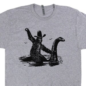Puede incluir: Una camiseta gris con una ilustraci&oacute;n en blanco y negro de un Bigfoot con un sombrero de vaquero montando un Nessie.