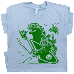 Puede incluir: Camiseta azul claro con un gr&aacute;fico verde de Godzilla con gafas de sol y una tabla de surf. El dise&ntilde;o incluye palmeras, olas y p&aacute;jaros. La tabla de surf tiene un s&iacute;mbolo de yin-yang.