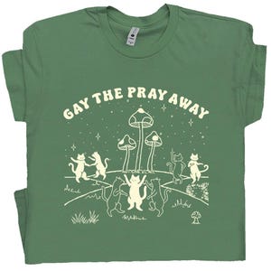 Puede incluir: Camiseta verde con el texto "GAY THE PRAY AWAY" sobre un diseño de gatos bailando, setas y estrellas. El diseño en blanco destaca sobre el fondo verde. Los gatos están en varias poses, y las setas tienen sombreros punteados.