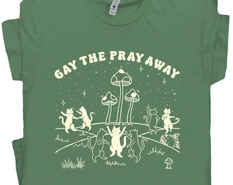 Puede incluir: Camiseta verde con el texto "GAY THE PRAY AWAY" sobre un diseño de gatos bailando, setas y estrellas. El diseño en blanco destaca sobre el fondo verde. Los gatos están en varias poses, y las setas tienen sombreros punteados.