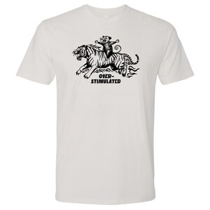 May include: White t-shirt featuring a black and white graphic of a tiger being ridden by a small animal. The text "OVER-STIMULATED" is printed below the tiger. The shirt is made of a soft, comfortable material.