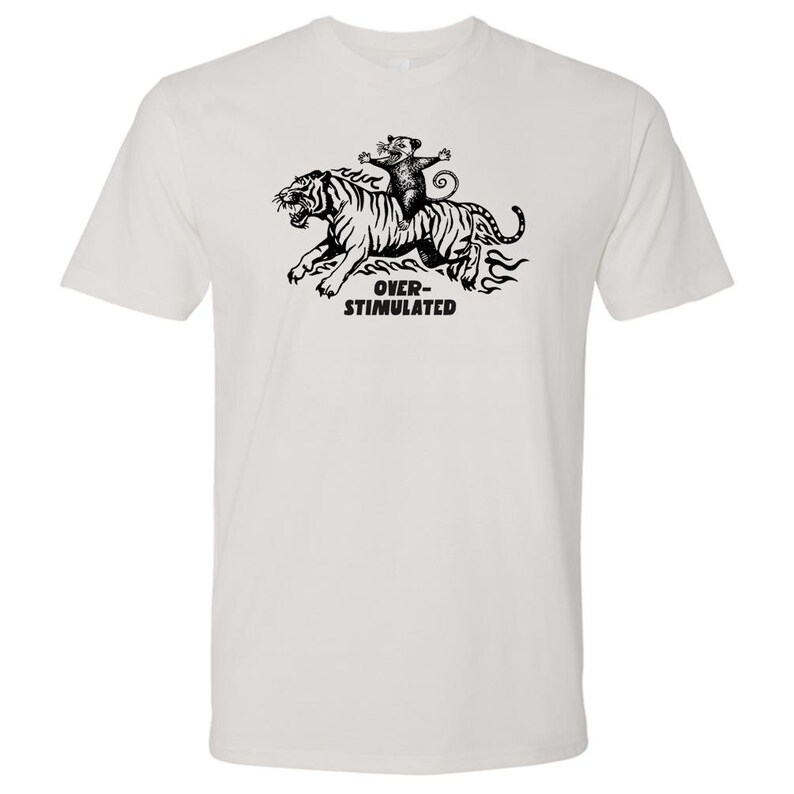 May include: White t-shirt featuring a black and white graphic of a tiger being ridden by a small animal. The text "OVER-STIMULATED" is printed below the tiger. The shirt is made of a soft, comfortable material.