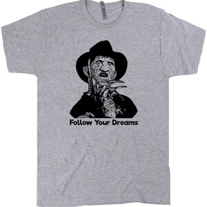 Op de afbeelding: Een grijs T-shirt met een zwart-wit afbeelding van Freddy Krueger uit de film A Nightmare on Elm Street. De tekst "Follow Your Dreams" is onder de afbeelding gedrukt.