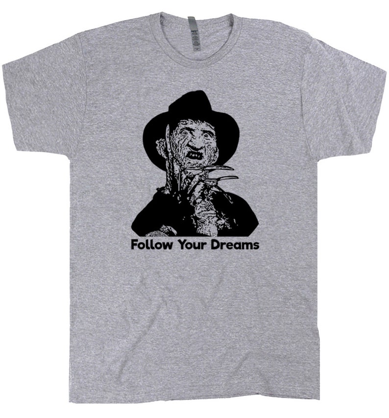 Op de afbeelding: Een grijs T-shirt met een zwart-wit afbeelding van Freddy Krueger uit de film A Nightmare on Elm Street. De tekst "Follow Your Dreams" is onder de afbeelding gedrukt.