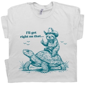 May include: White t-shirt with a teal graphic of a sloth wearing a cowboy hat riding a turtle. The sloth gives a thumbs-up, and the text reads "I'll get right on that..."