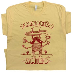 May include: A light yellow t-shirt with a cartoon illustration of a mustached insect wearing a sombrero, holding a beer and a taco. The words "TRANQUILO" and "AMIGO" are printed in red.