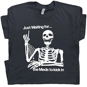 Könnte beinhalten: Dunkelgraues T-Shirt mit weißer Skelettgrafik. Das Skelett zeigt nach oben, mit dem Text "Just Waiting for..." darüber und "the Meds to kick in" darunter. Das Design ist eine humorvolle Interpretation des Wartens auf Medikamente.
