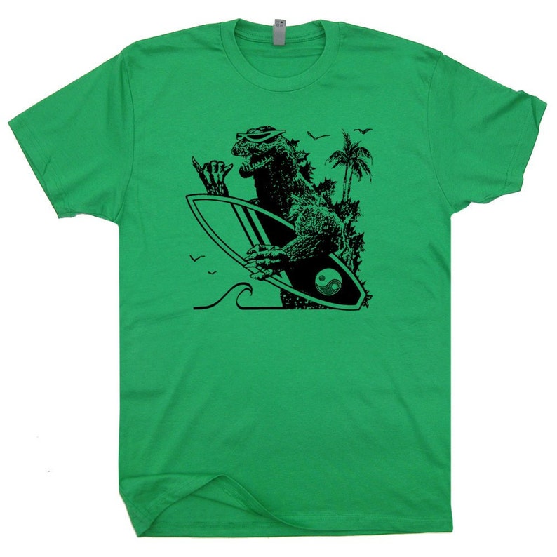 May include: Green t-shirt with a black and white graphic of a monster surfing. The monster is wearing sunglasses and has a yin yang symbol on its surfboard.