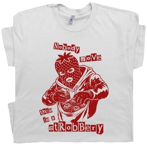 May include: White t-shirt featuring a red graphic of a person in a strawberry balaclava, holding strawberries. The text reads "Nobody move" and "this is a stRobbery".