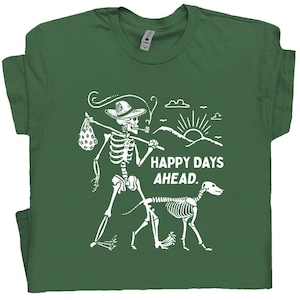 May include: Green t-shirt with a white graphic of a skeleton with a dog, a hat, and a bindle. The text "HAPPY DAYS AHEAD" is printed in white. The skeleton is smoking a pipe and walking with the dog.
