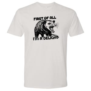 May include: White t-shirt featuring a black and white illustration of an opossum with the text "FIRST OF ALL I'M A DELIGHT". The opossum is depicted with its mouth open, showing its teeth.