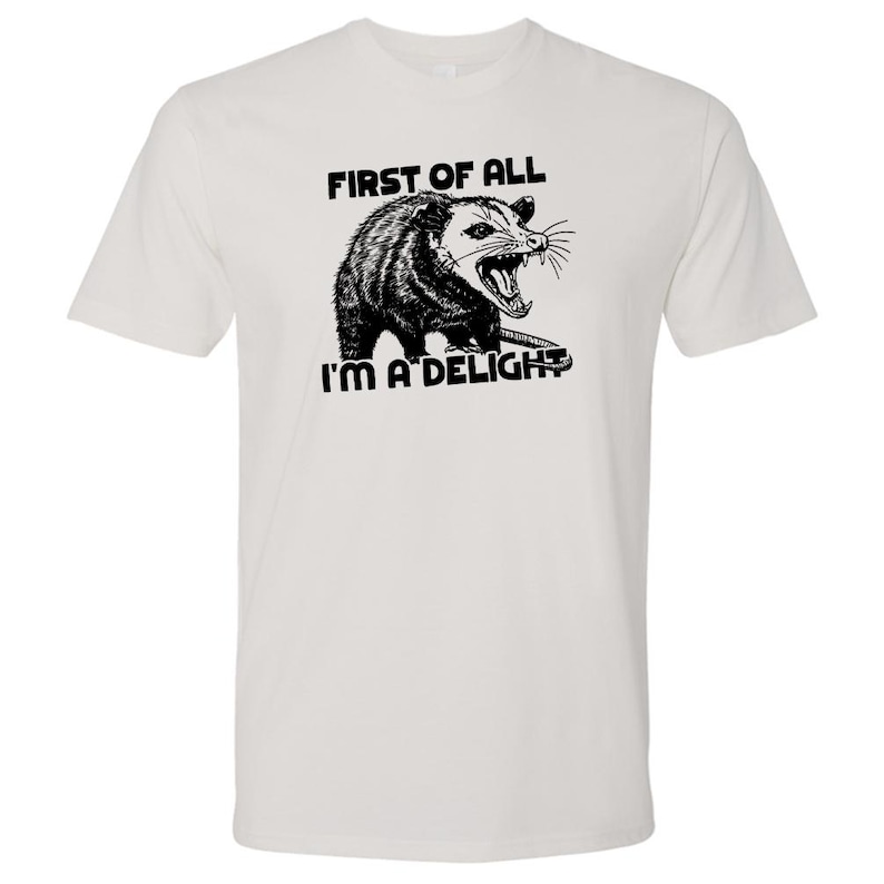 May include: White t-shirt featuring a black and white illustration of an opossum with the text "FIRST OF ALL I'M A DELIGHT". The opossum is depicted with its mouth open, showing its teeth.