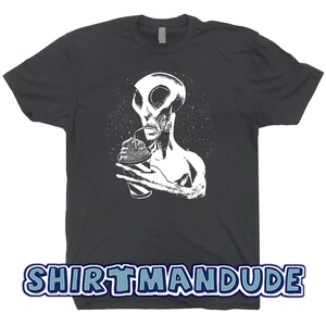 May include: A dark gray t-shirt with a white graphic of an alien drinking from a cup with a straw. The alien has large eyes and a pointed head. The background is a dark gray with a white starry pattern. The text "SHIRTMANDUDE" is printed in blue on the bottom of the shirt.