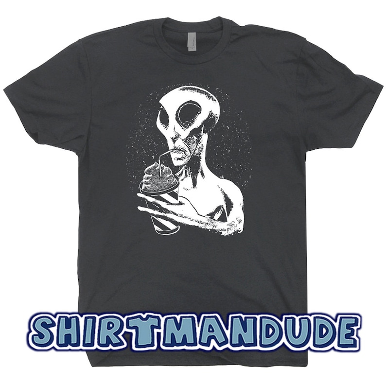 May include: A dark gray t-shirt with a white graphic of an alien drinking from a cup with a straw. The alien has large eyes and a pointed head. The background is a dark gray with a white starry pattern. The text "SHIRTMANDUDE" is printed in blue on the bottom of the shirt.