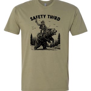 May include: Olive green t-shirt with a black graphic of a cowboy riding a bear, with the text "SAFETY THIRD" above the image. The cowboy is holding a bottle in the air. The design includes trees and birds.