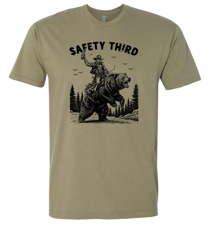 May include: Olive green t-shirt with a black graphic of a cowboy riding a bear, with the text "SAFETY THIRD" above the image. The cowboy is holding a bottle in the air. The design includes trees and birds.