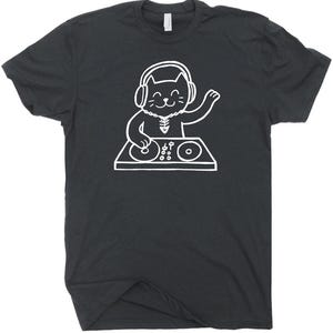 May include: Dark gray t-shirt featuring a white illustration of a cat DJ. The cat is wearing headphones and a necklace, with one paw raised. The cat is positioned behind a DJ mixing board with two turntables and various controls. The shirt has a crew neck.