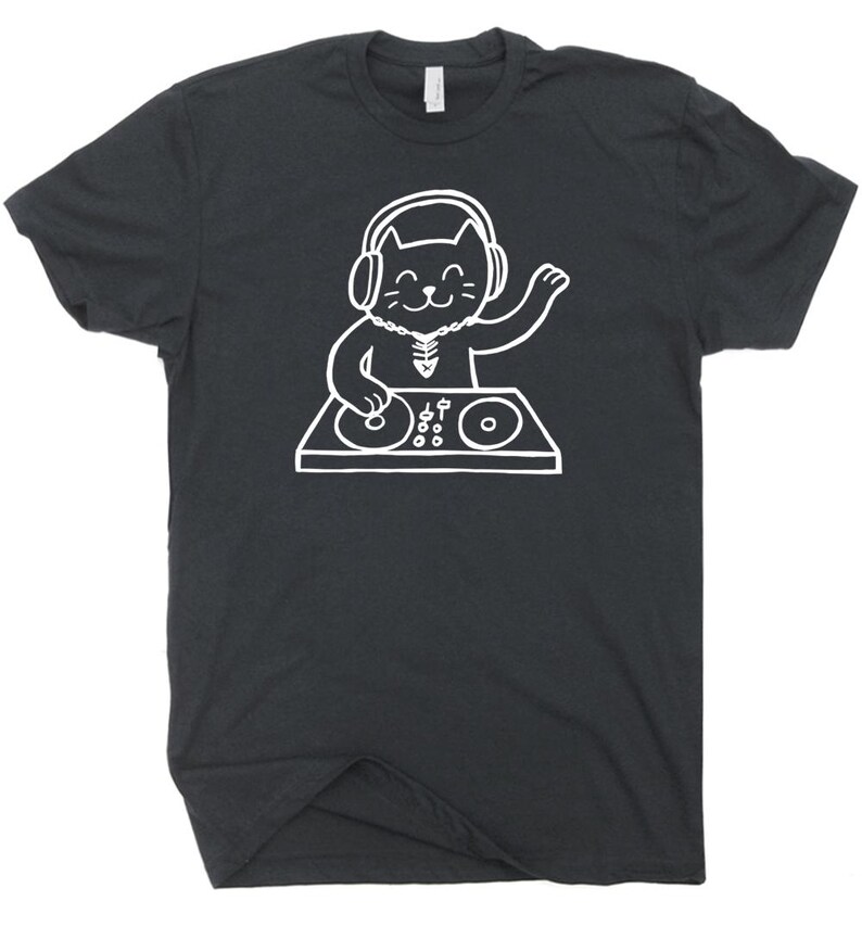 May include: Dark gray t-shirt featuring a white illustration of a cat DJ. The cat is wearing headphones and a necklace, with one paw raised. The cat is positioned behind a DJ mixing board with two turntables and various controls. The shirt has a crew neck.