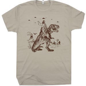 May include: A tan t-shirt featuring a brown graphic of a figure riding a Tyrannosaurus Rex. The graphic includes a volcano, flying saucers, and a Bigfoot-like figure. The shirt is made of a soft material and has a crew neck.