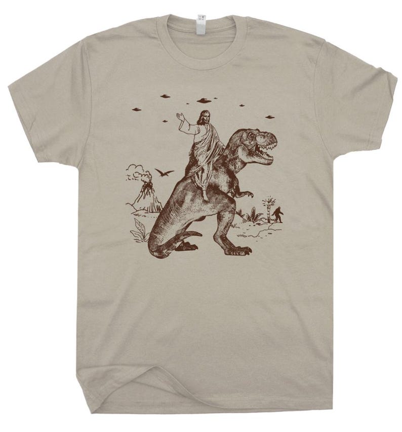May include: A tan t-shirt featuring a brown graphic of a figure riding a Tyrannosaurus Rex. The graphic includes a volcano, flying saucers, and a Bigfoot-like figure. The shirt is made of a soft material and has a crew neck.