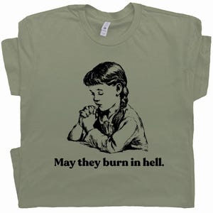 以下が含まれることがあります： オリーブグリーンのTシャツ。祈る少女の白黒イラストがプリントされています。画像の下には「May they burn in hell」のテキスト。カジュアルウェアです。