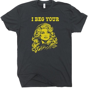 May include: Dark grey t-shirt featuring a yellow illustration of a woman's portrait and the text "I BEG YOUR" above the portrait. The woman has long, wavy hair and large hoop earrings. The shirt has a crew neck.