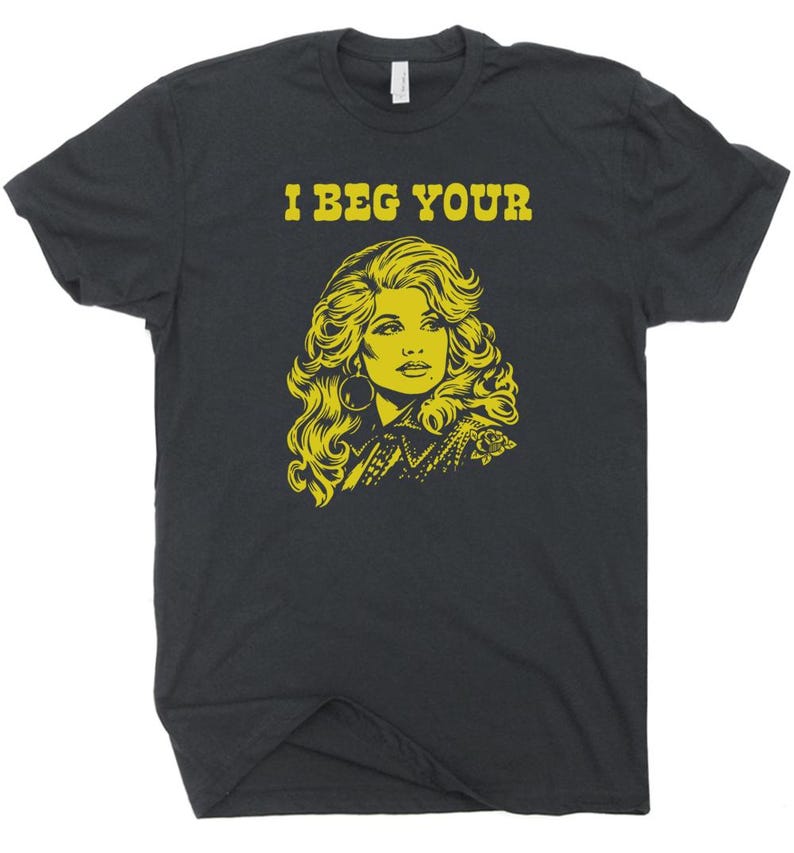 May include: Dark grey t-shirt featuring a yellow illustration of a woman's portrait and the text "I BEG YOUR" above the portrait. The woman has long, wavy hair and large hoop earrings. The shirt has a crew neck.