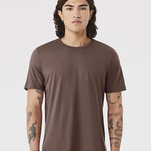 May include: A solid brown crew neck t-shirt. The short-sleeved shirt is made of a soft material. The person wearing the shirt has tattoos on their arms.