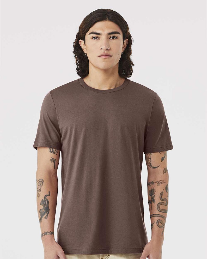 May include: A solid brown crew neck t-shirt. The short-sleeved shirt is made of a soft material. The person wearing the shirt has tattoos on their arms.