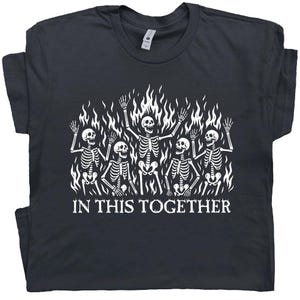 May include: Dark gray t-shirt featuring a white graphic of six skeletons in front of flames, with their arms raised. The text "IN THIS TOGETHER" is printed below the skeletons. The shirt is made of a soft material.