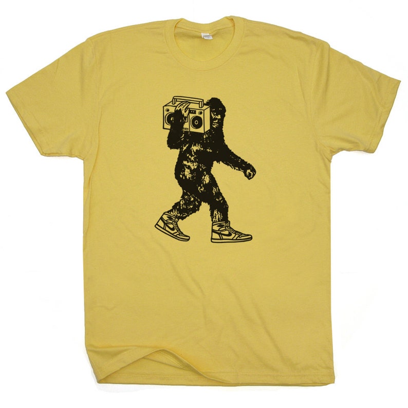 Puede incluir: Una camiseta amarilla con un gr&aacute;fico negro de un Bigfoot llevando una radio de cassette en el hombro. El Bigfoot lleva zapatillas deportivas.