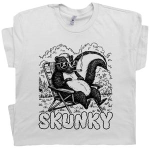 May include: White t-shirt featuring a relaxed skunk wearing sunglasses and smoking a cigar while sitting in a folding chair. The word "SKUNKY" is printed in large, bubble letters below the illustration.