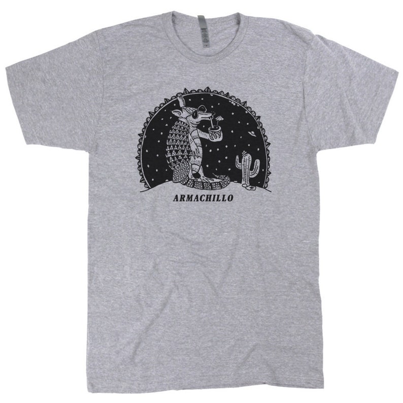May include: A gray t-shirt with a black and white illustration of an armadillo wearing sunglasses and drinking from a glass. The armadillo is standing in front of a cactus and the text "ARMACHILLO" is printed below the illustration.