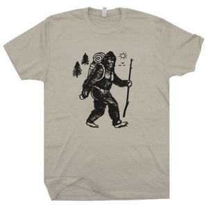 May include: A beige t-shirt with a black graphic of a Bigfoot-like creature carrying a backpack and walking with a stick. The graphic also includes trees, a sun, and birds.