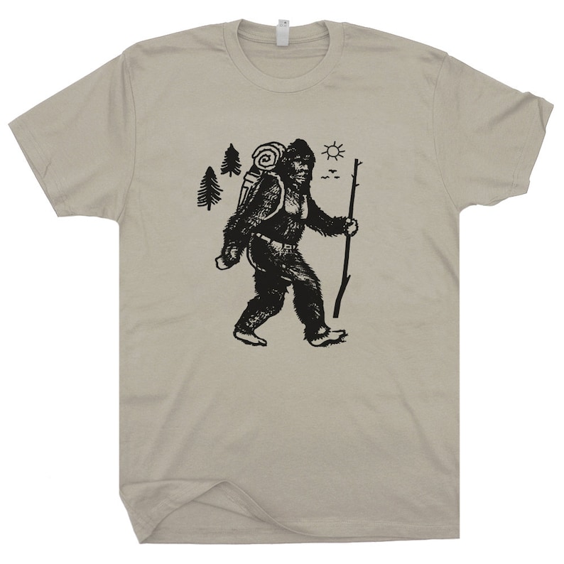 May include: A beige t-shirt with a black graphic of a Bigfoot-like creature carrying a backpack and walking with a stick. The graphic also includes trees, a sun, and birds.