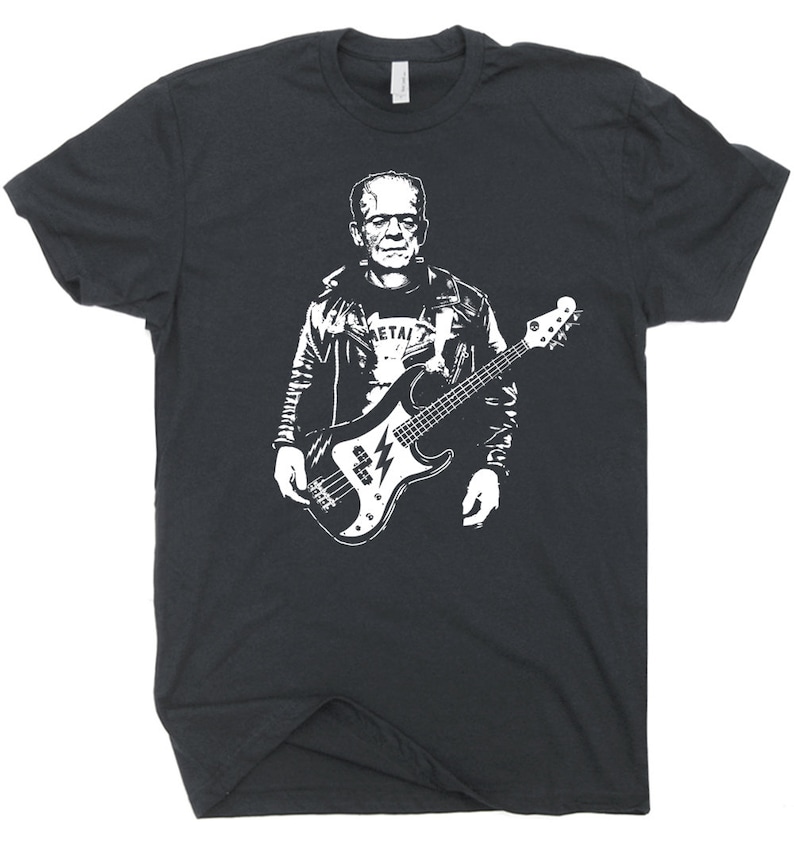 May include: Dark gray t-shirt featuring a white graphic of Frankenstein playing a bass guitar. The design includes a leather jacket and a shirt that says "METAL". The bass guitar has a lightning bolt design.