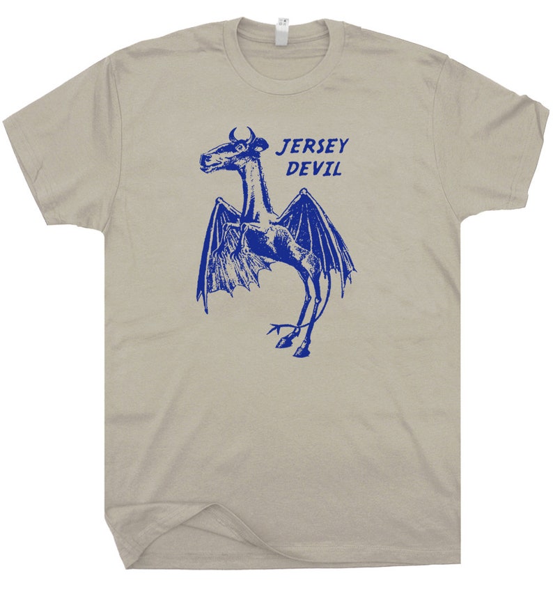 May include: A light gray t-shirt with a blue graphic of the Jersey Devil, a mythical creature with wings, a horse-like head, and a long tail. The text "JERSEY DEVIL" is printed in blue above the creature.