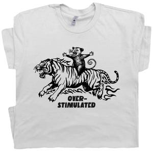 May include: White t-shirt with a black graphic of a tiger and a possum. The possum is riding the tiger, and the text "OVER-STIMULATED" is below the image. The shirt has a crew neck and short sleeves.