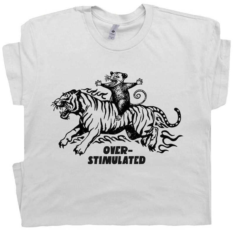 May include: White t-shirt with a black graphic of a tiger and a possum. The possum is riding the tiger, and the text "OVER-STIMULATED" is below the image. The shirt has a crew neck and short sleeves.