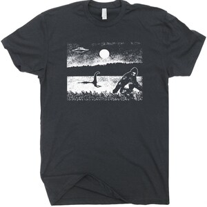 Puede incluir: Una camiseta negra con un estampado gr&aacute;fico blanco de un OVNI, Bigfoot y el monstruo del Lago Ness. El gr&aacute;fico est&aacute; en un estilo desgastado.