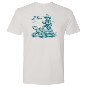 May include: White t-shirt with a teal graphic of a sloth wearing a cowboy hat riding a turtle. The text "I'll get right on that..." is above the illustration. The sloth gives a thumbs up.