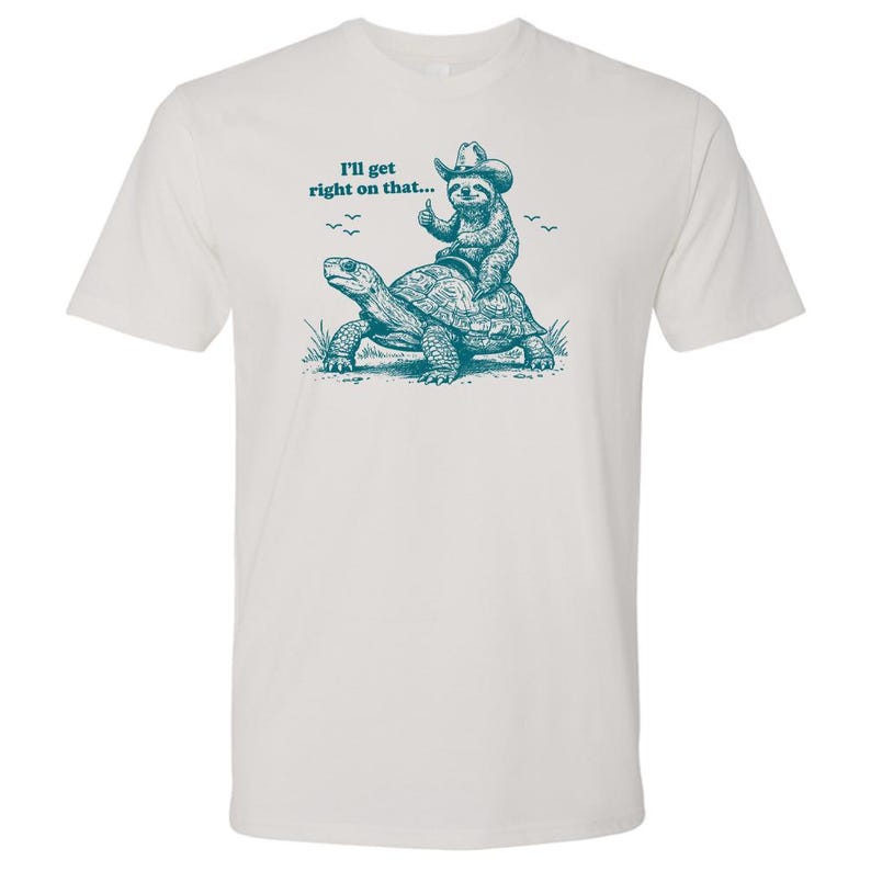May include: White t-shirt with a teal graphic of a sloth wearing a cowboy hat riding a turtle. The text "I'll get right on that..." is above the illustration. The sloth gives a thumbs up.