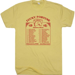 May include: A yellow t-shirt with a red graphic of a Chinese restaurant menu. The menu includes a variety of dishes, such as chicken chow mein, pepper steak with onion, and pork egg foo young. The text "Lucky Fortune" is at the top of the menu, and the text "Chinatown NYC 212-864-2121" is at the bottom.