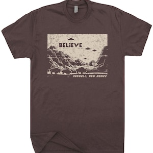 May include: A brown t-shirt with a vintage-style graphic of a mountain landscape with the words "BELIEVE" and "ROSWELL, NEW MEXICO" printed on it. There are several flying saucers in the sky above the mountains.