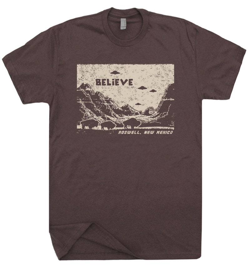 May include: A brown t-shirt with a vintage-style graphic of a mountain landscape with the words "BELIEVE" and "ROSWELL, NEW MEXICO" printed on it. There are several flying saucers in the sky above the mountains.