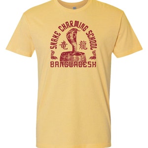 Pode incluir: Camiseta amarela com um gr&aacute;fico vermelho de um encantador de cobras com o texto "Snake Charming School Bangladesh" em um c&iacute;rculo.