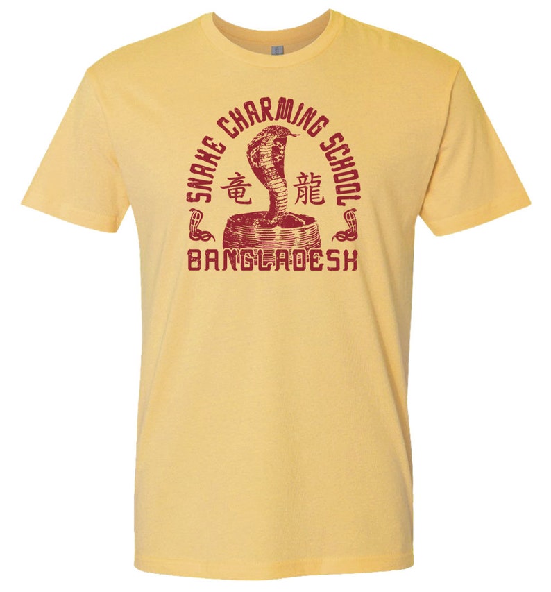 Pode incluir: Camiseta amarela com um gr&aacute;fico vermelho de um encantador de cobras com o texto "Snake Charming School Bangladesh" em um c&iacute;rculo.