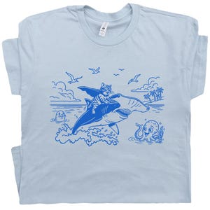 May include: Light blue t-shirt featuring a blue illustration of a cat riding a shark through the ocean. The design includes an octopus, a seal, birds, and palm trees. The cat is wearing a cape and glasses.