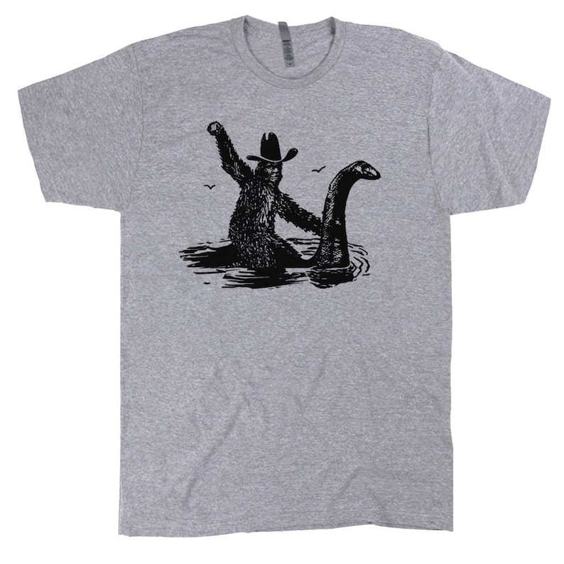 Puede incluir: Una camiseta gris con una ilustraci&oacute;n en blanco y negro de un Bigfoot con un sombrero de vaquero montando un Nessie.