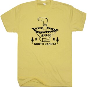 Puede incluir: Una camiseta amarilla con un gr&aacute;fico negro de un calcet&iacute;n de dibujos animados en un cono de tr&aacute;fico con el texto "FARGO Est. 1871 NORTH DAKOTA"